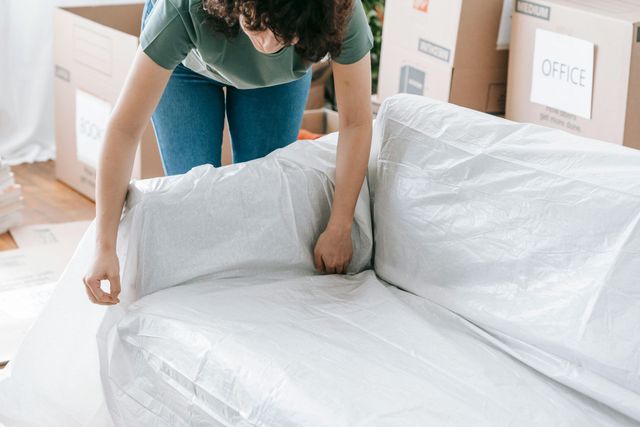 Woman packing a white sofa in plastic wrap during a move, surrounded by cardboard boxes.