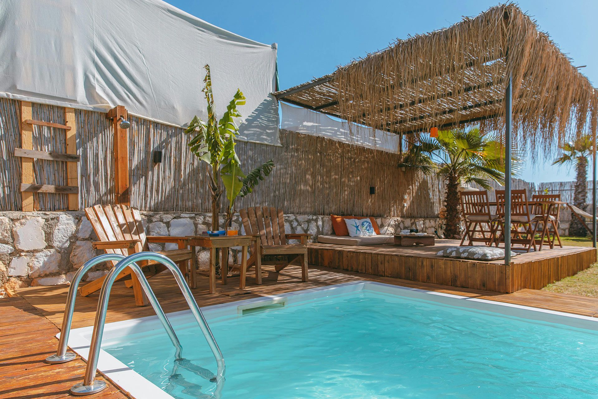Poolside patio with turquoise pool, wicker chairs, and a shaded thatched pergola beside a white building
