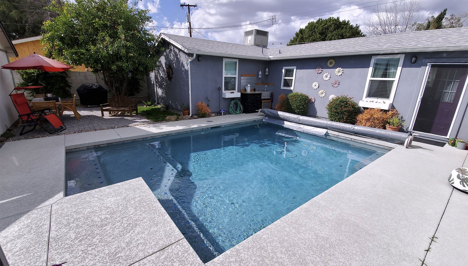 Backyard swimming pool beside a gray house, with patio chairs and a red umbrella