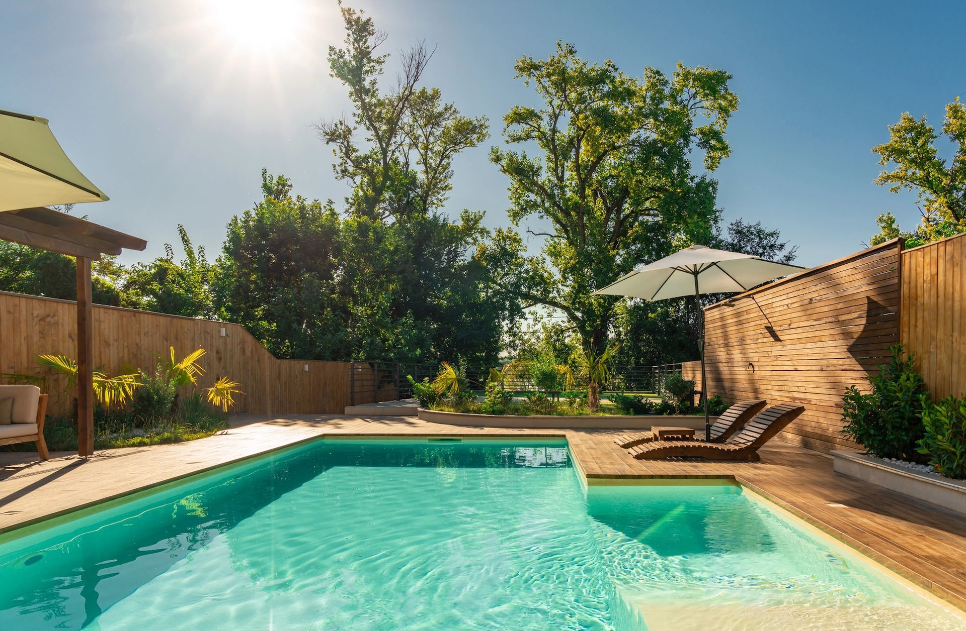Sunny backyard pool with lounge chairs, umbrellas, and a wooden privacy fence