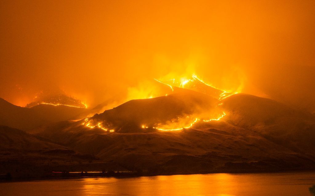 Montañas ardiendo por la noche; las llamas encienden las laderas, reflejándose en el agua de abajo.