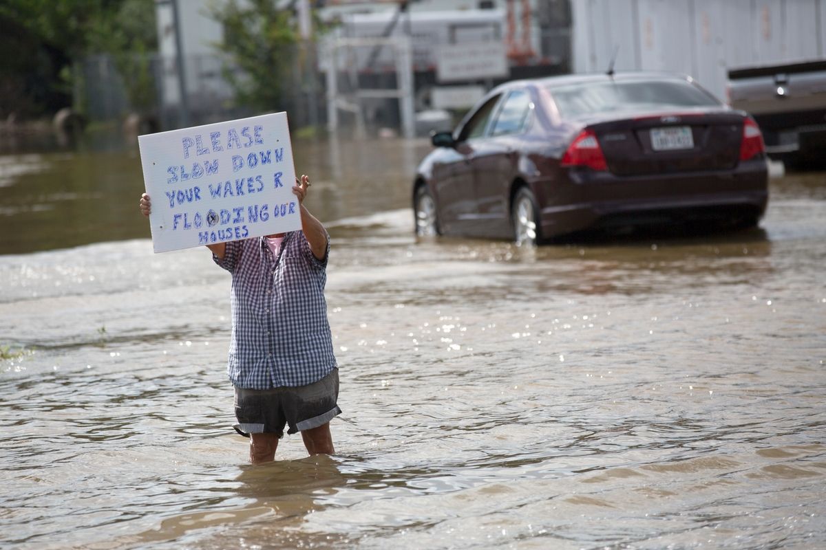 Person wading through floodwater holding a protest sign, with a submerged car in the background.