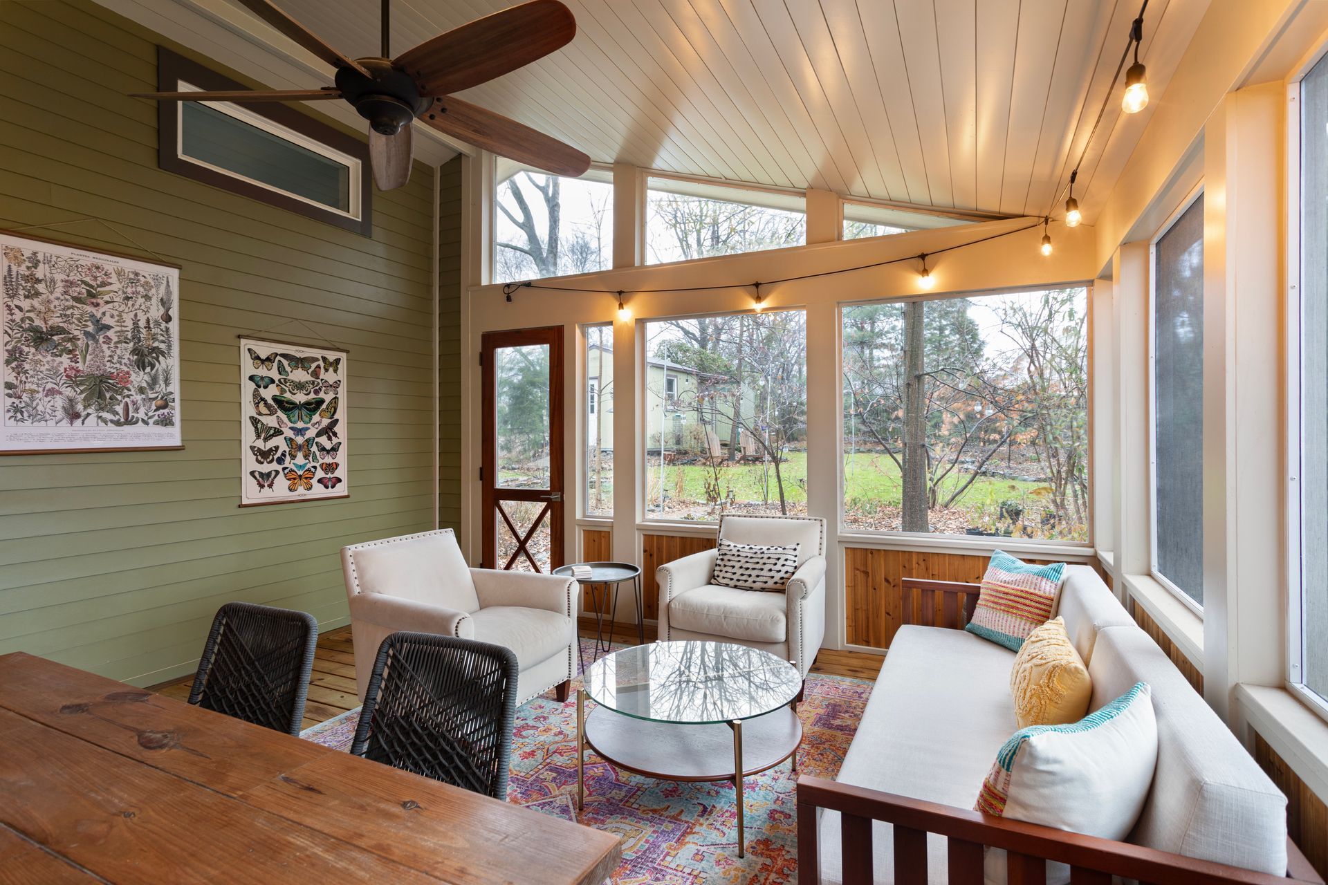 Bright sunroom with white sofas, wooden table, and large windows overlooking trees