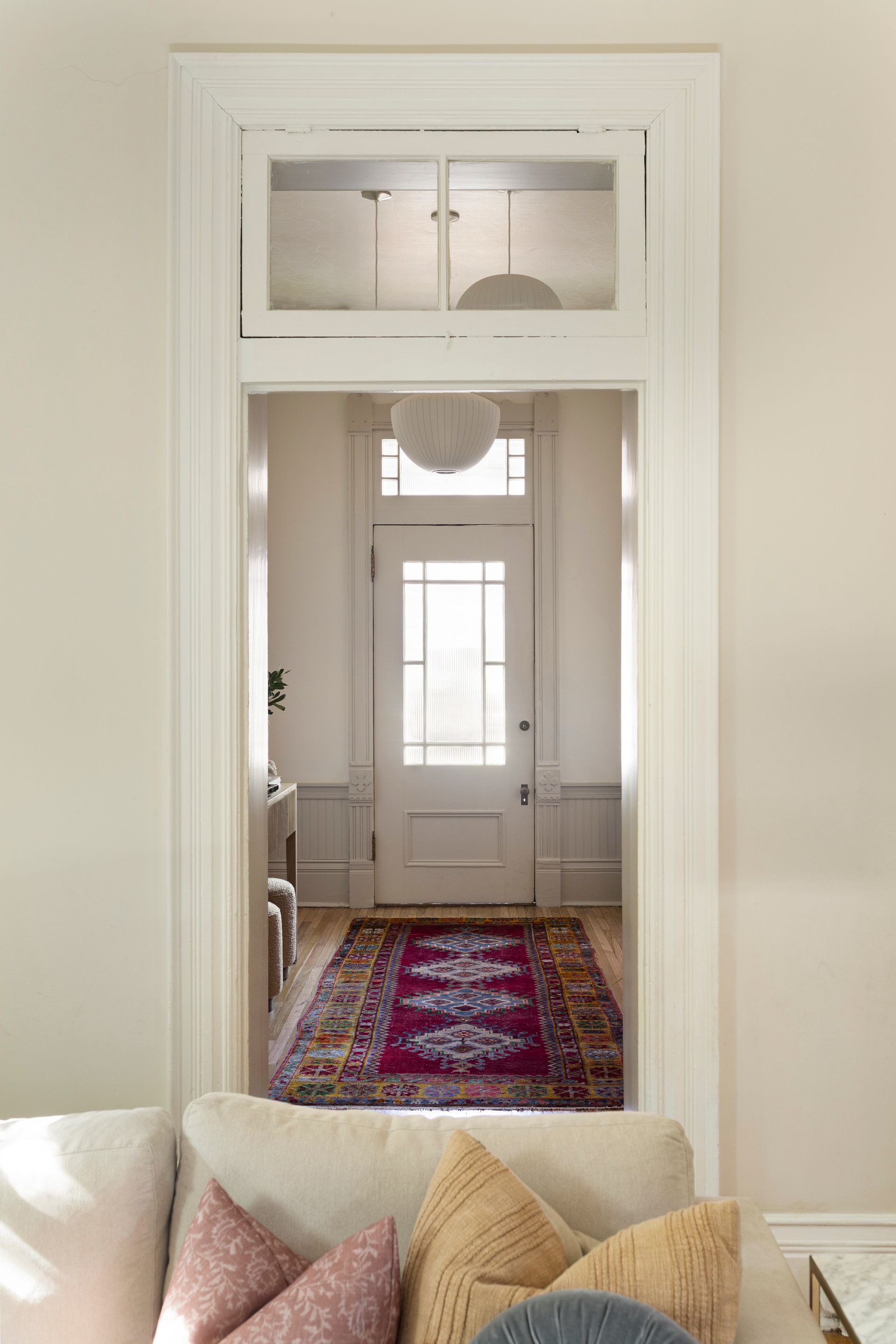 Bright hallway view through a white doorway, with a patterned red rug and front door visible beyond a sofa.