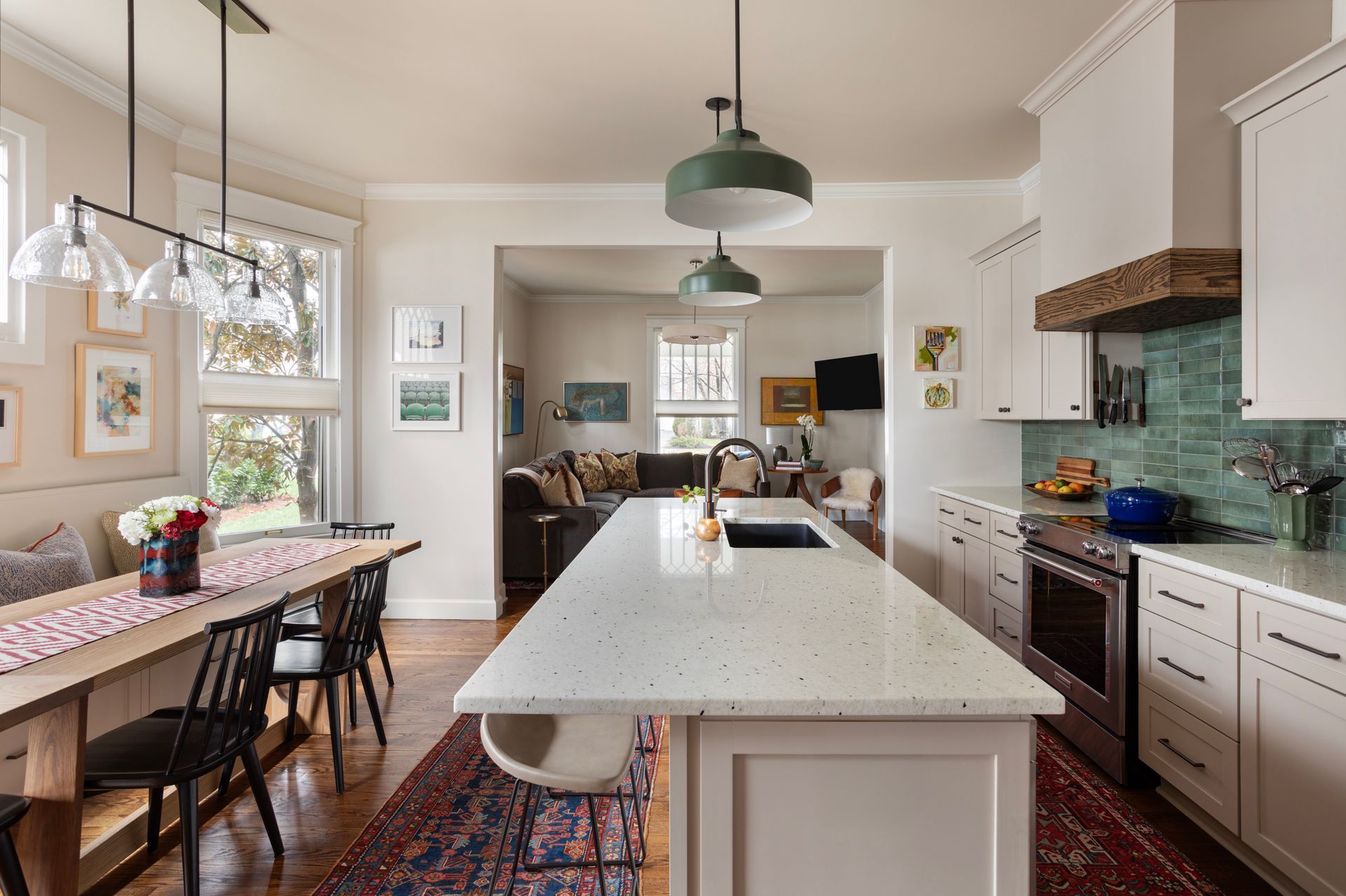 Bright kitchen with white island, green pendant lights, teal backsplash, and dining table by windows