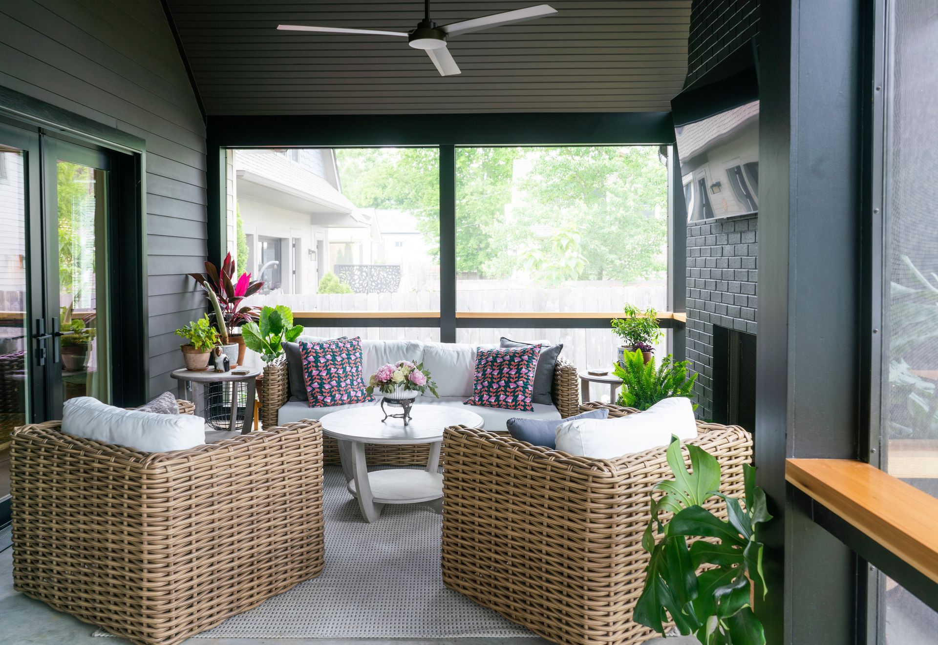 Covered porch with wicker seating, white cushions, colorful pillows, and a ceiling fan overlooking a bright yard