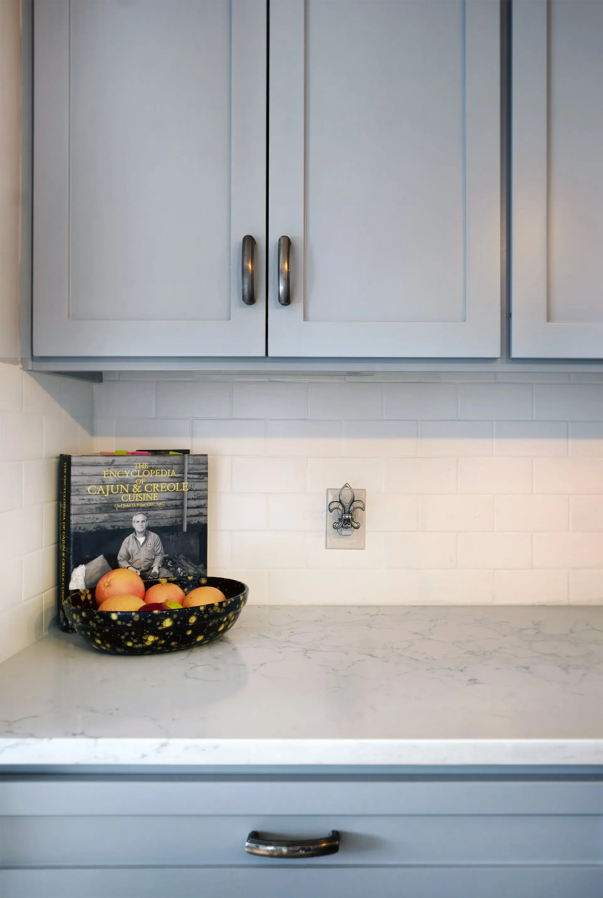 Light gray kitchen cabinets above a marble countertop with a fruit basket and cookbook on the counter.