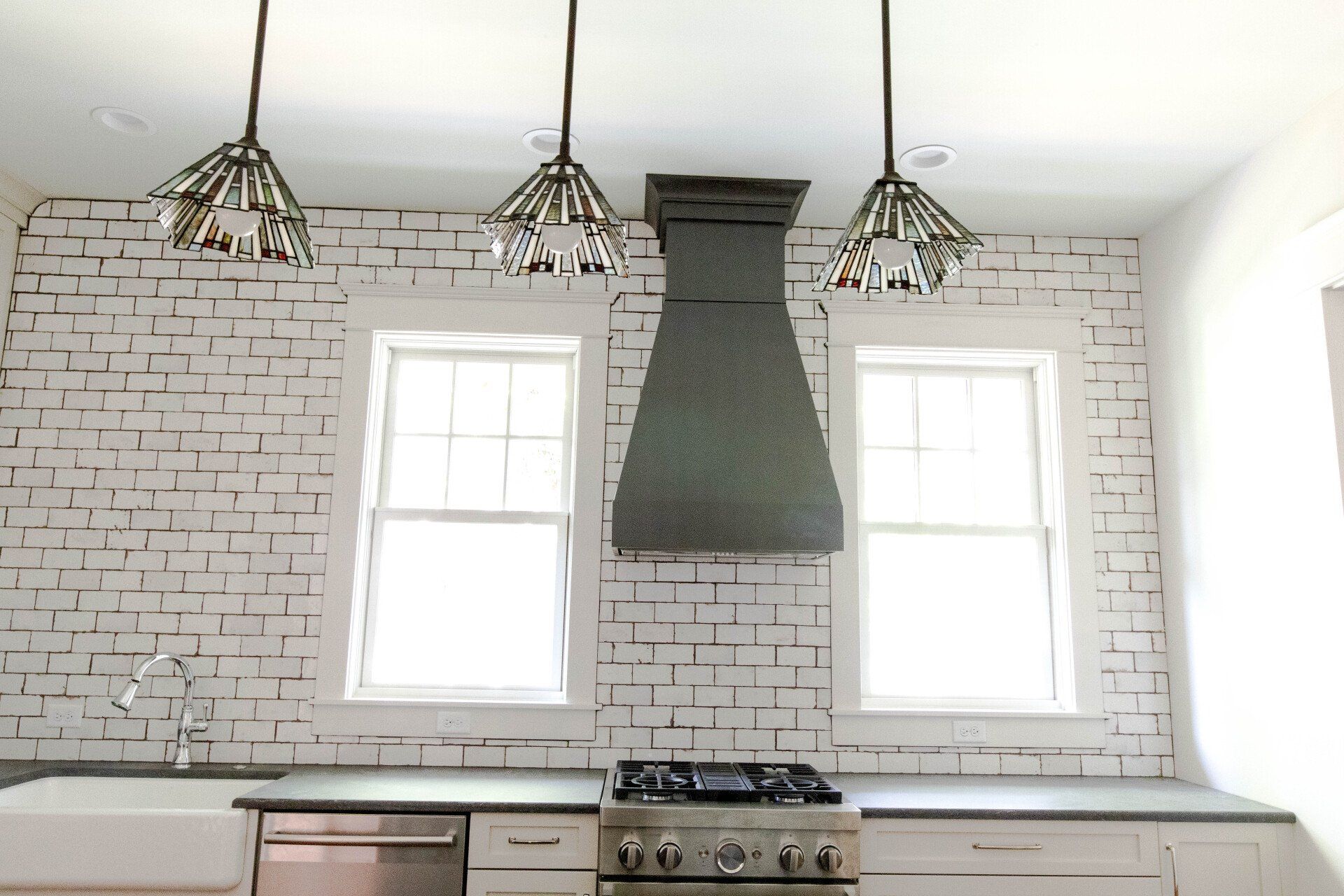 Modern kitchen with white brick backsplash, two windows, dark range hood, and three pendant lights