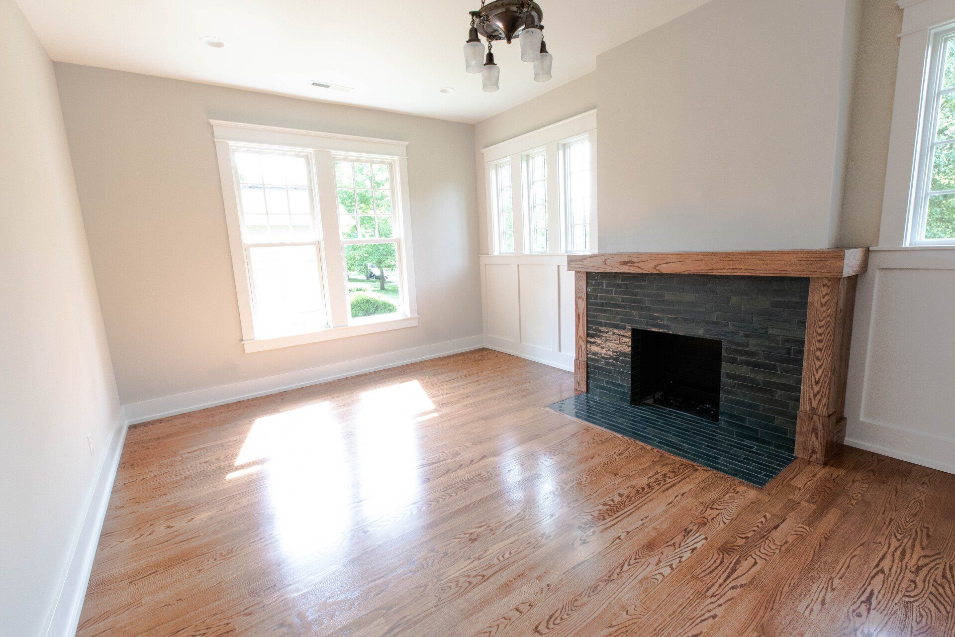 Empty living room with hardwood floors, large windows, and a brick fireplace.