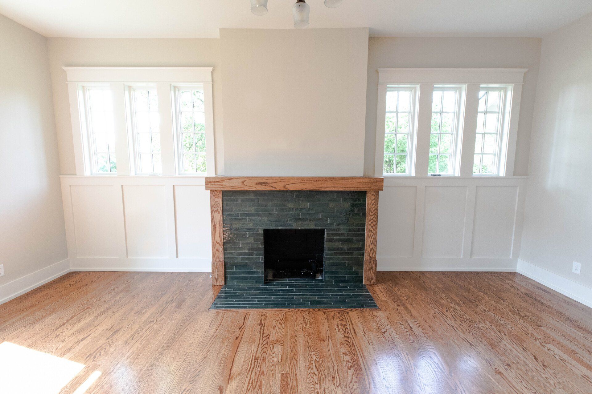 Empty living room with hardwood floors, white walls, and a central brick fireplace flanked by windows.