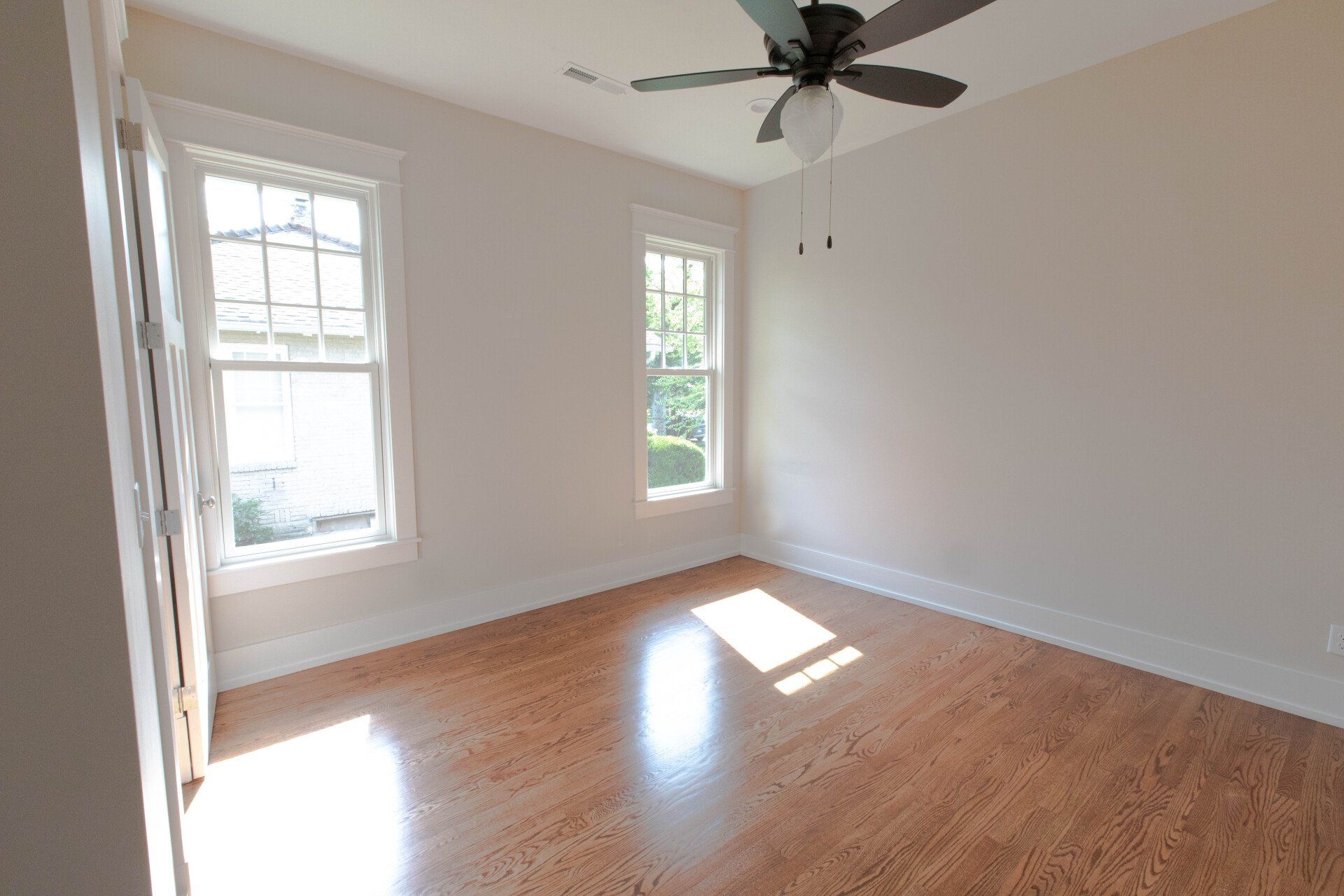 Empty sunlit room with hardwood floors, two windows, and a ceiling fan
