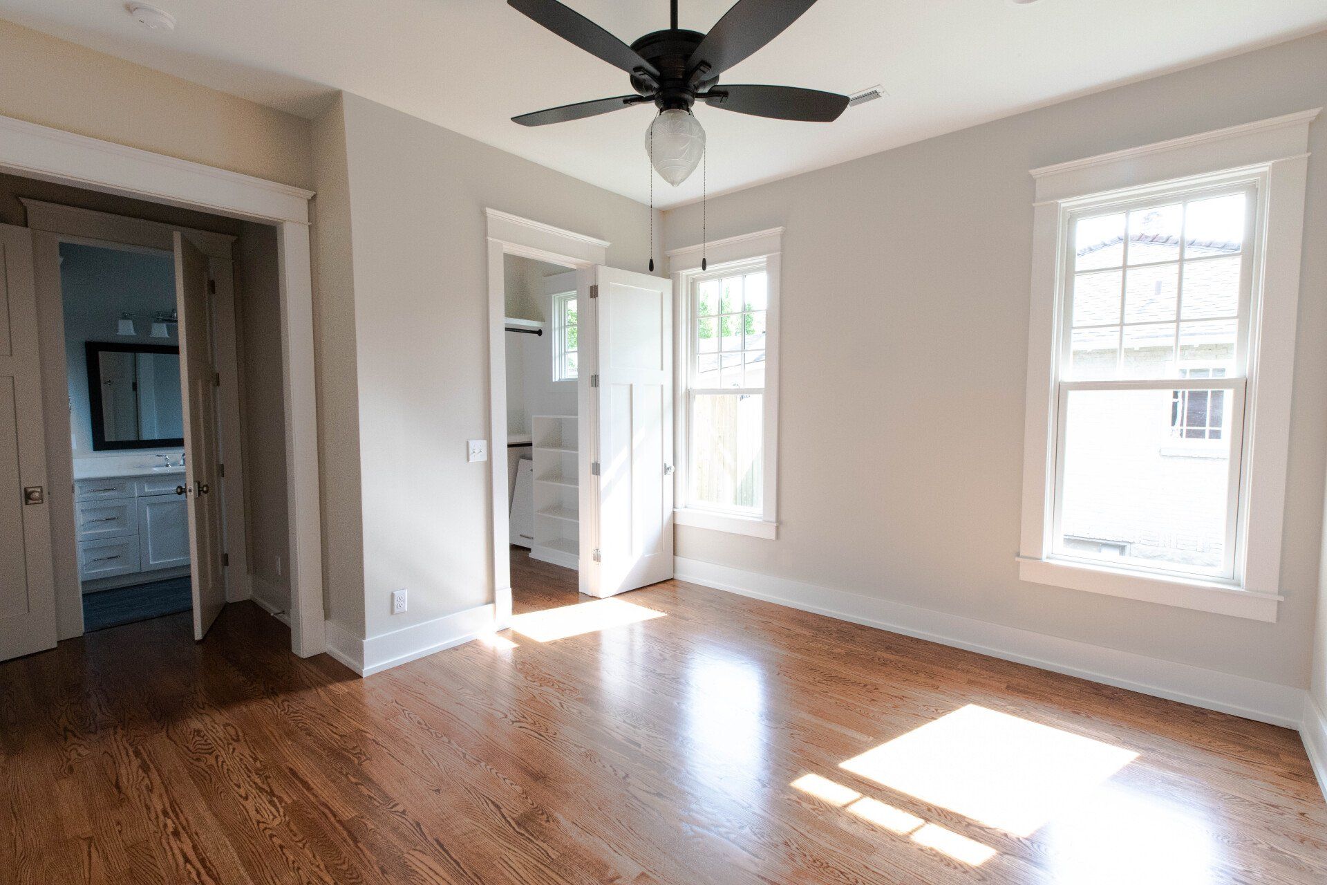 Empty sunlit room with hardwood floors, white walls, two windows, and a ceiling fan