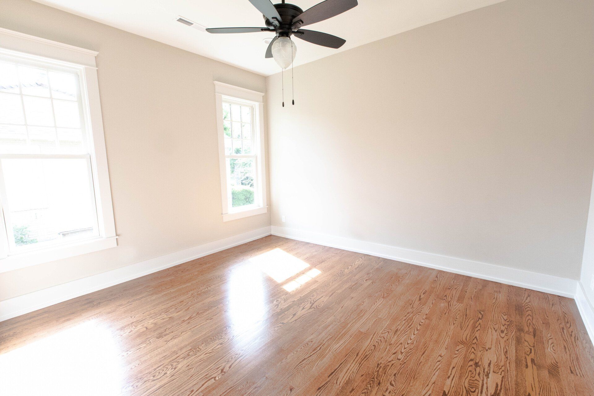 Empty bright room with hardwood floors, white walls, two windows, and a ceiling fan