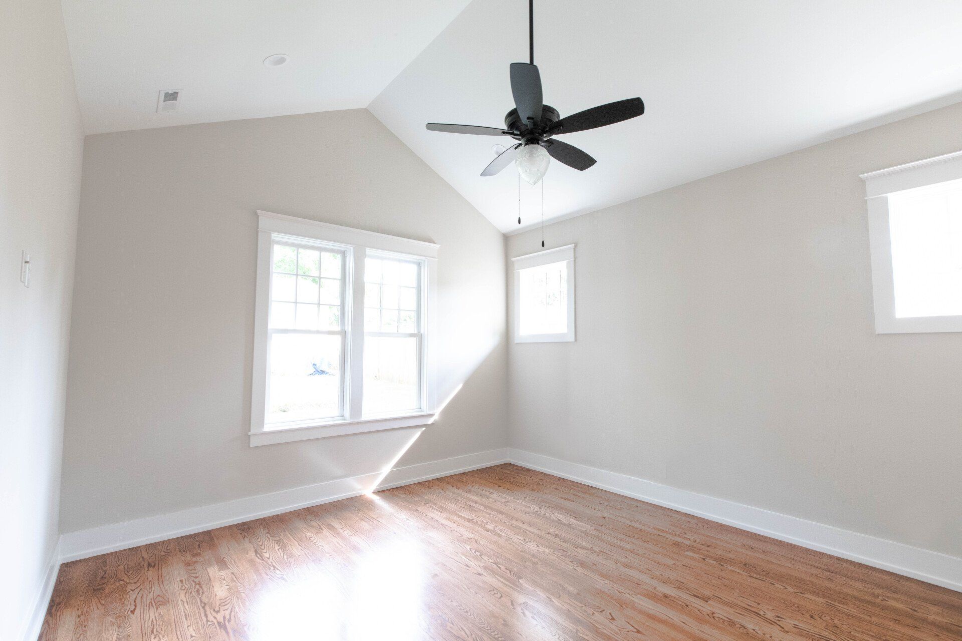 Empty white bedroom with hardwood floors, vaulted ceiling, black ceiling fan, and three windows