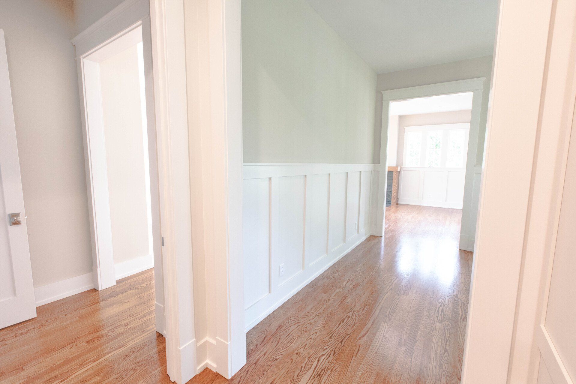 Bright hallway with white wainscoting, hardwood floors, and open doorways leading to a sunlit room.