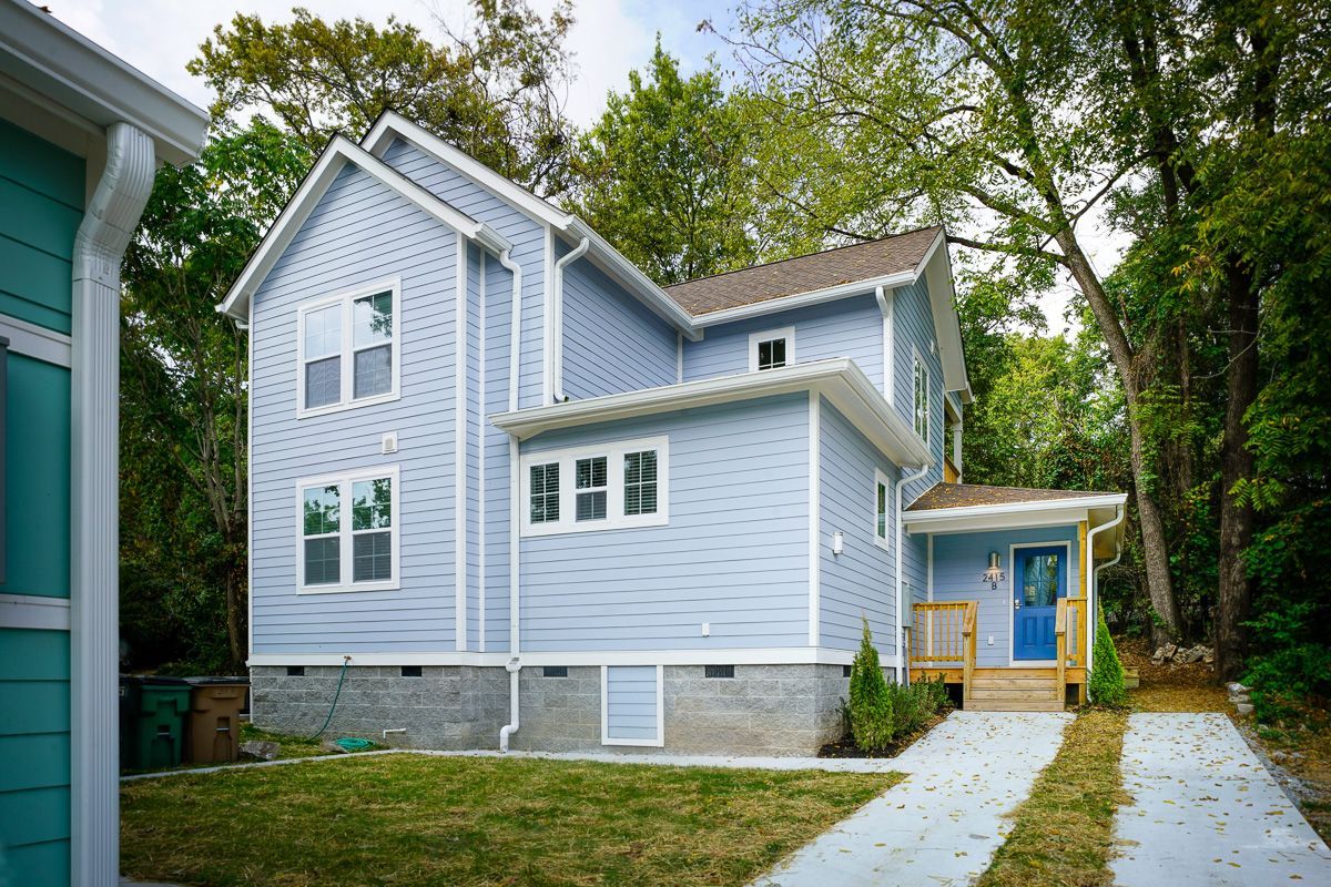 Blue house with white trim, stone foundation, and a driveway beside tall trees