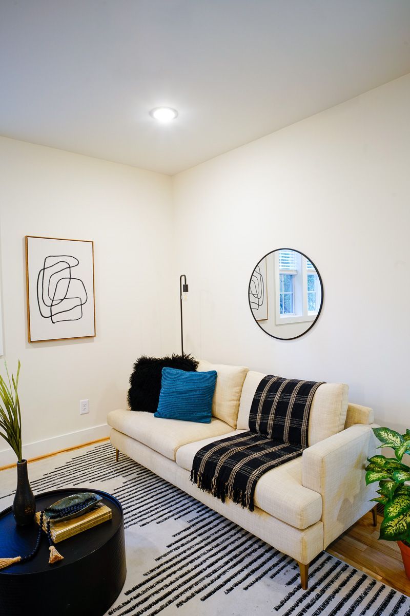 Cozy white living room with a cream sofa, blue pillows, striped rug, wall art, and round mirror.