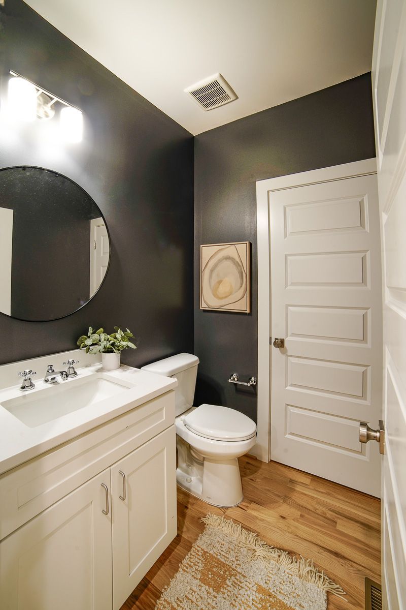 Small bathroom with dark walls, white vanity and toilet, round mirror, and wood floor.