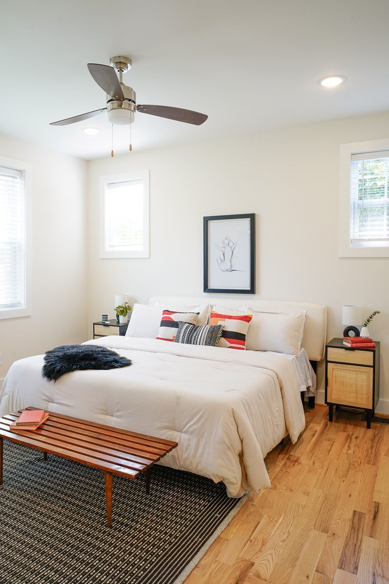 Bright bedroom with white bed, ceiling fan, wooden bench, and sunlit windows on hardwood floor