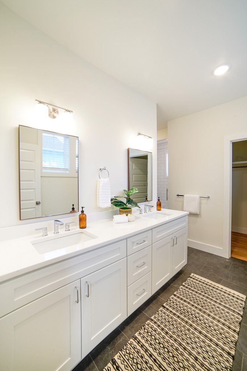 Bright bathroom with white double vanity, mirrors, and patterned rug on a tiled floor.