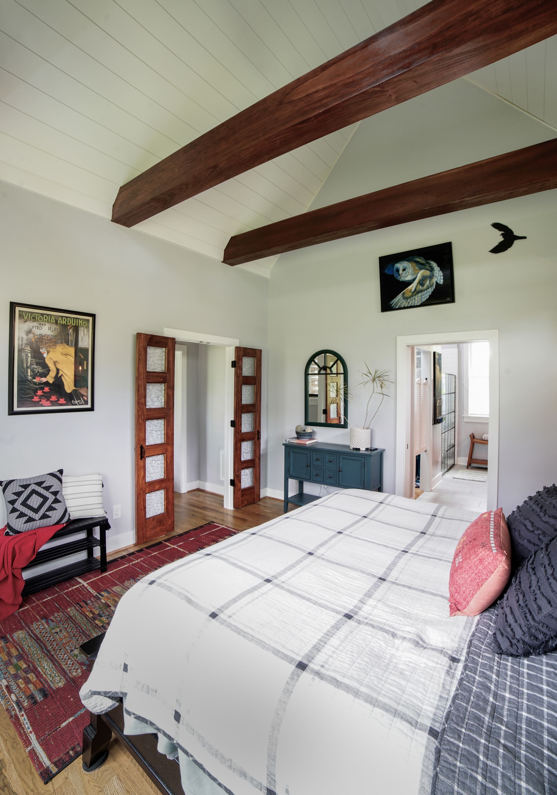 Cozy bedroom with white bedspread, exposed wood beams, red rug, and framed art on cream walls