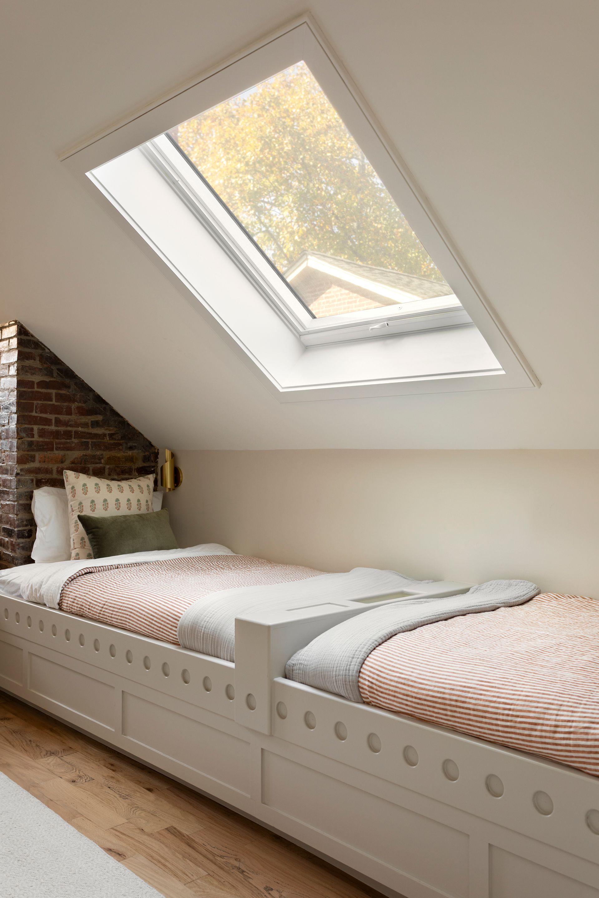 Loft bedroom with twin beds under a slanted skylight and exposed brick wall.