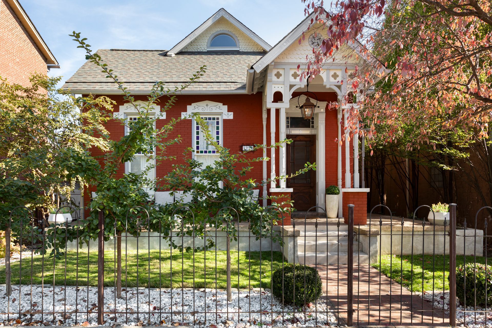 Red house with white trim and a front porch behind a black iron fence, framed by trees and a small garden.