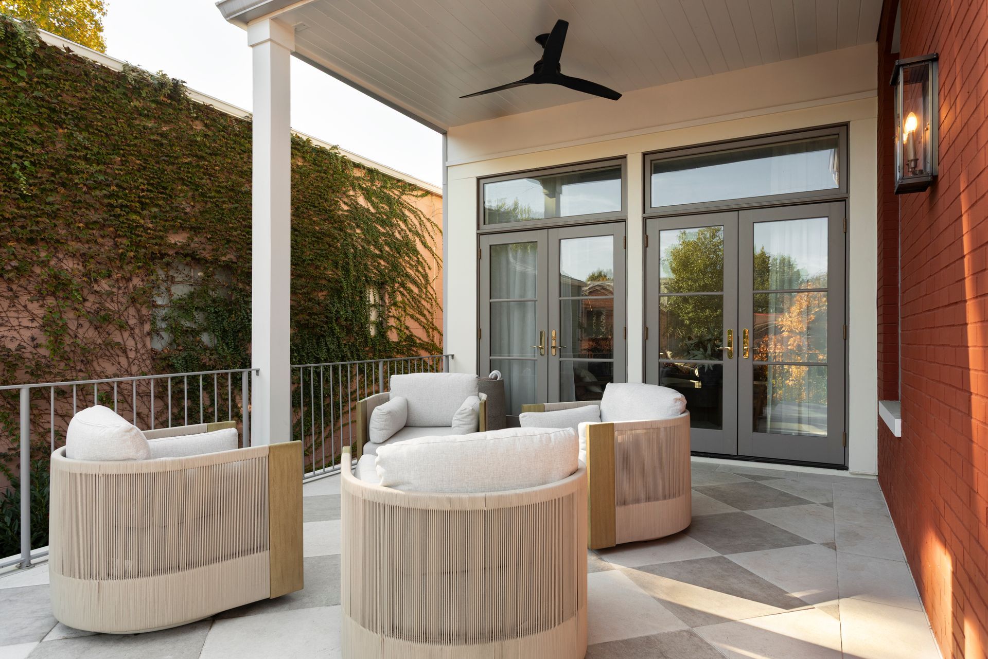 Covered patio with woven lounge chairs, round tables, and glass doors beside a brick wall