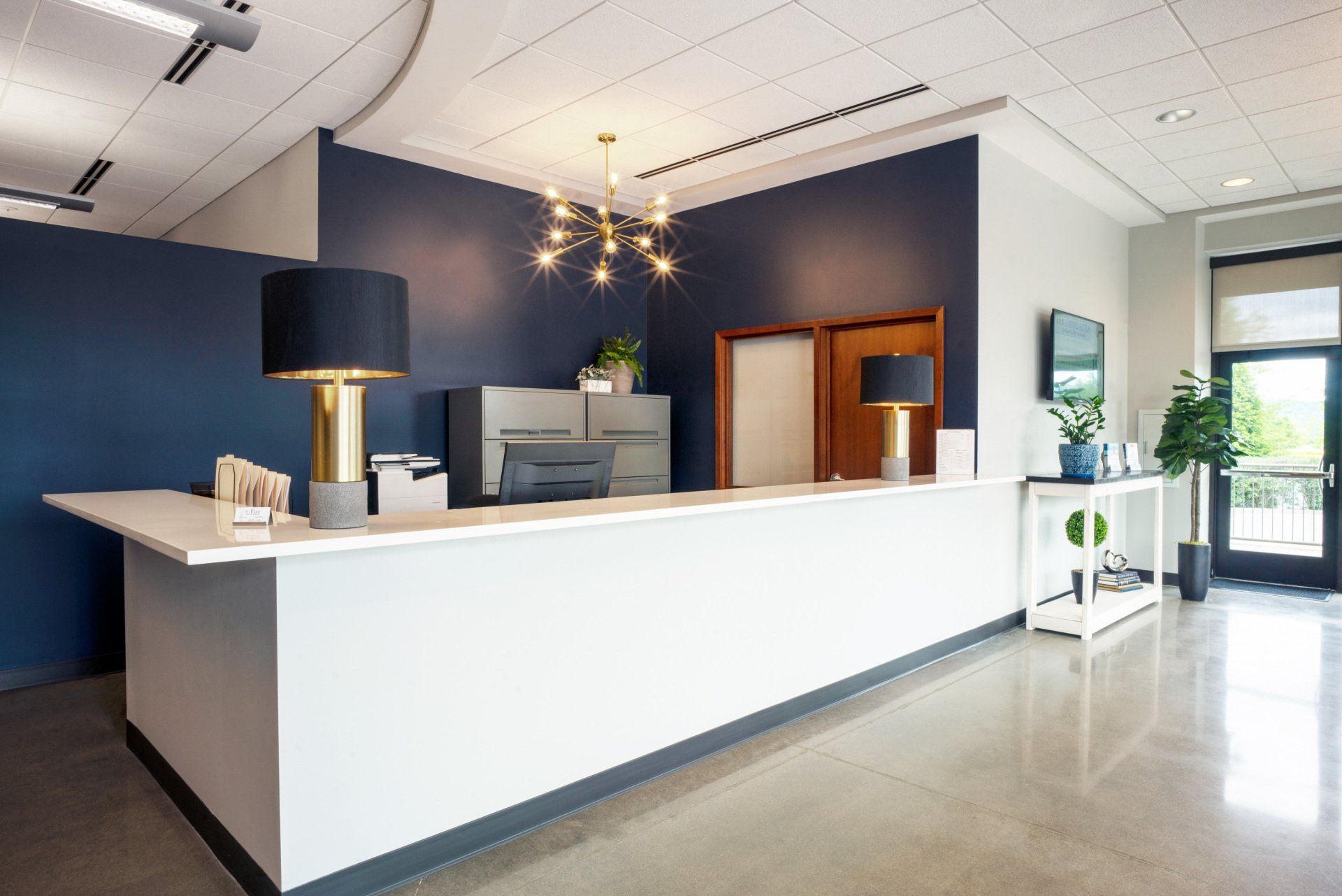 Modern hotel reception desk with white counter, dark blue walls, chandelier, and bright lobby entrance