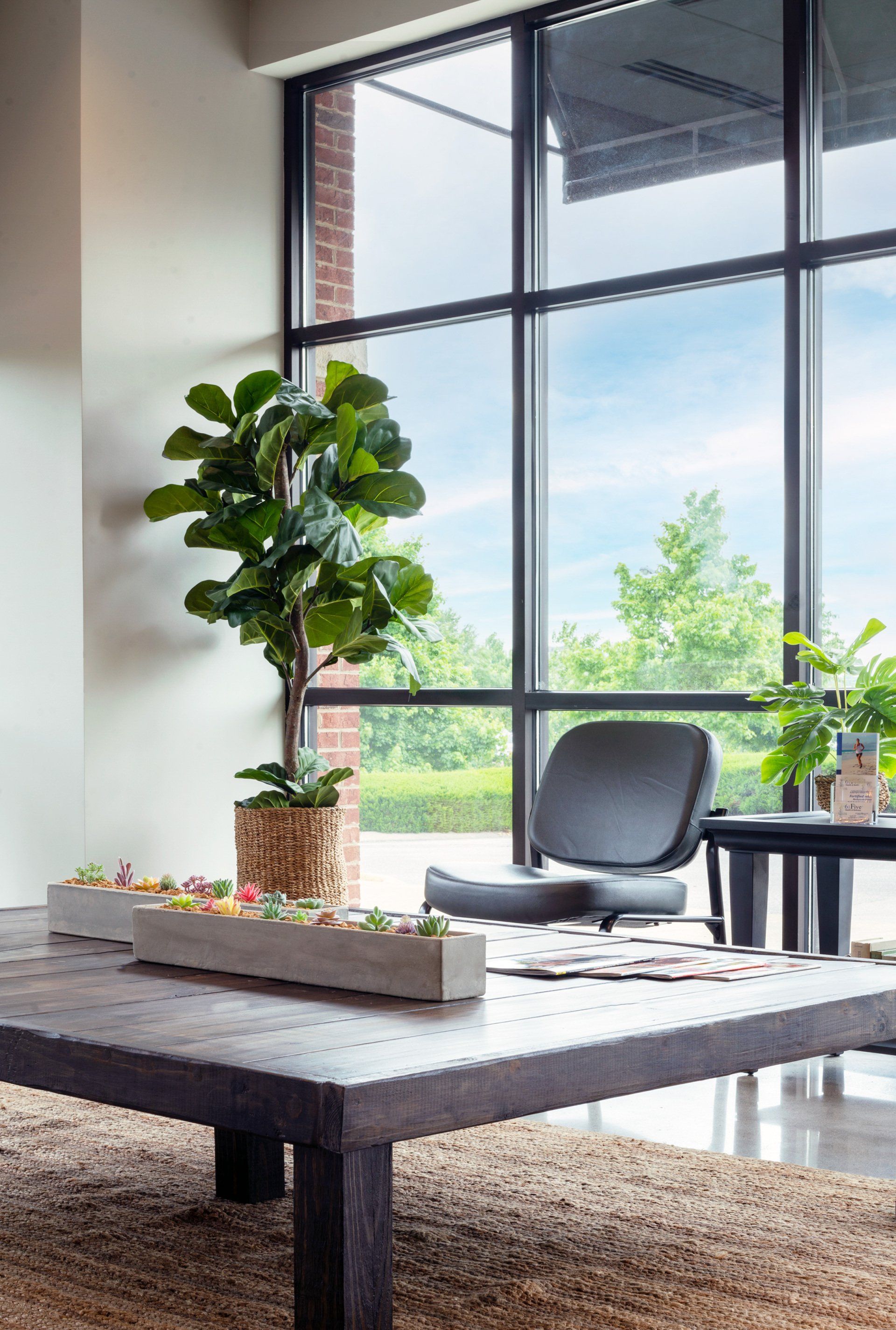 Modern living room with large windows, gray chair, potted plants, and a low coffee table by a city view