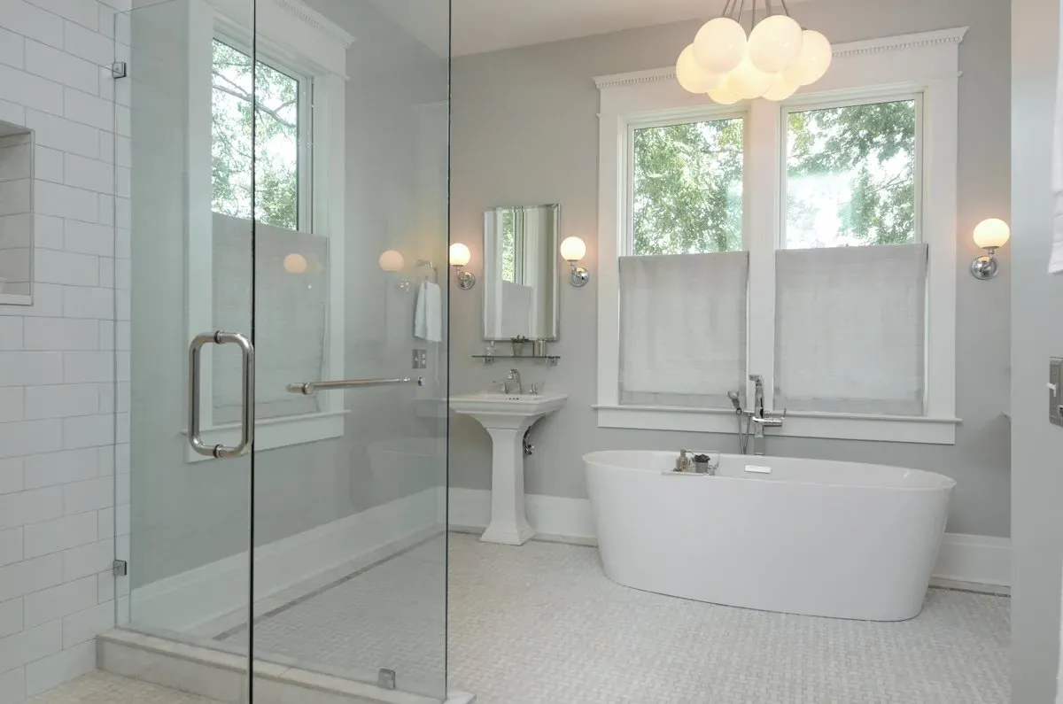 Bright white bathroom with glass shower, pedestal sink, clawfoot tub, and two windows with shades