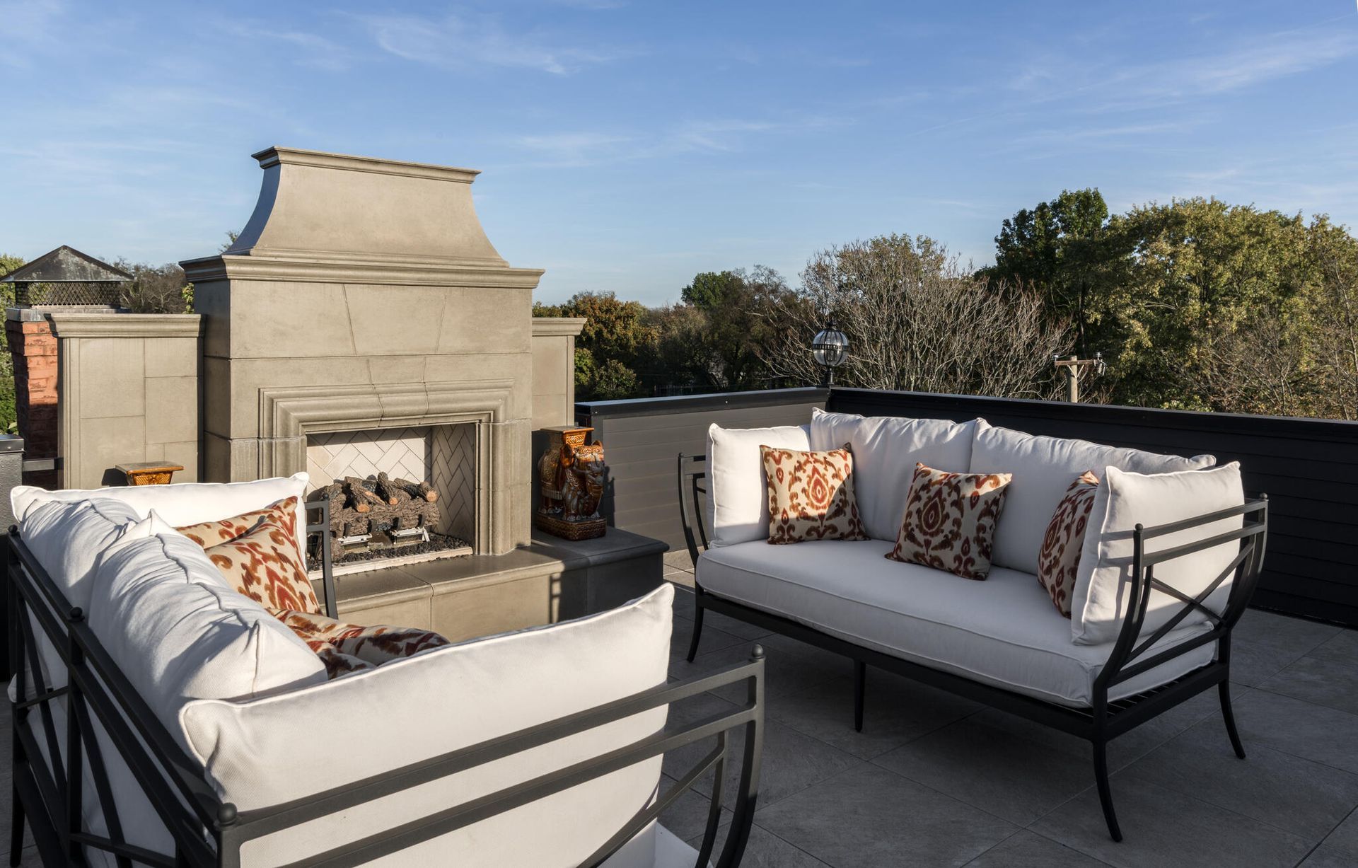 Patio seating area with white sofas and patterned pillows around a stone fireplace at sunset