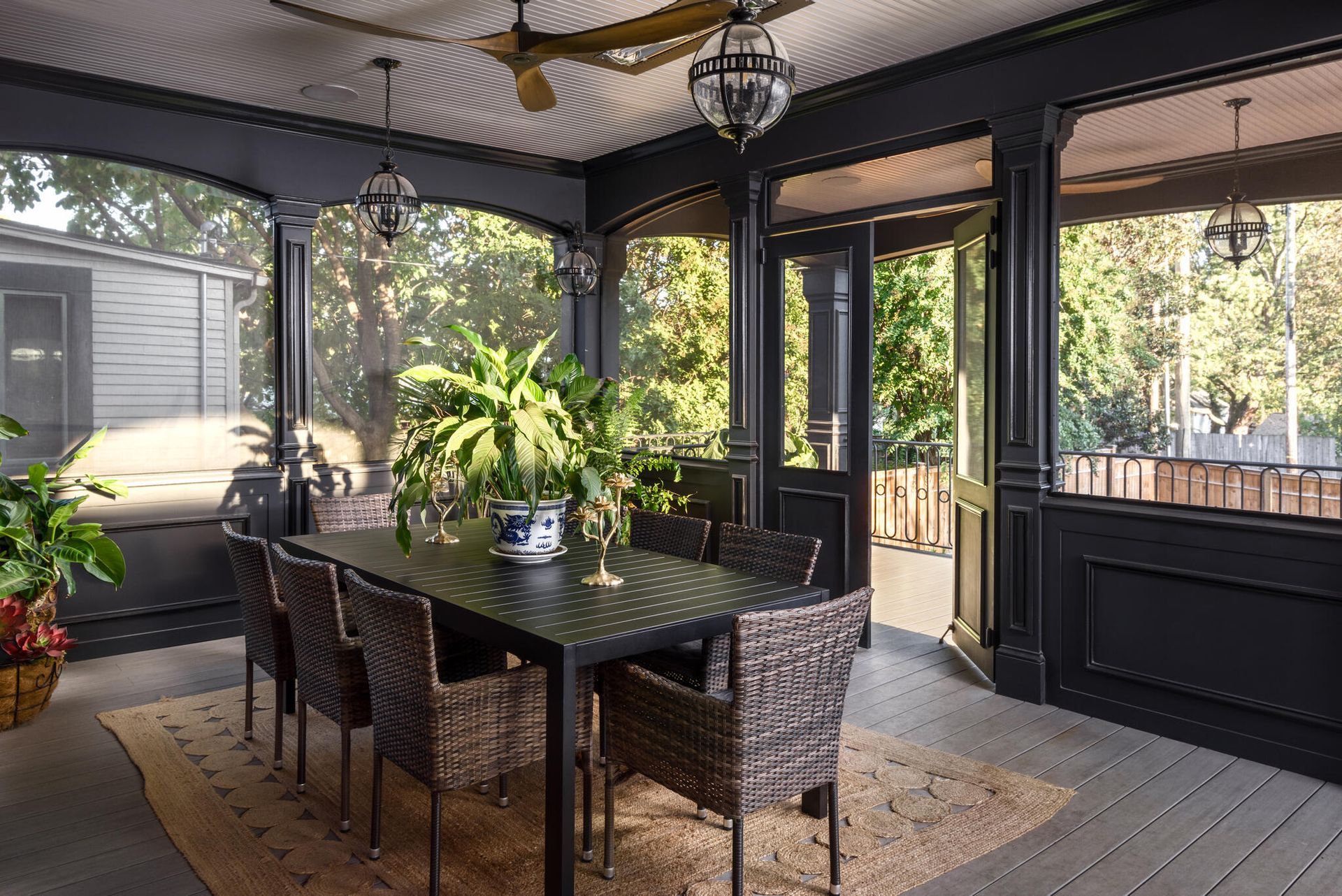 Covered patio dining area with wicker chairs, black table, potted plant, and large windows.