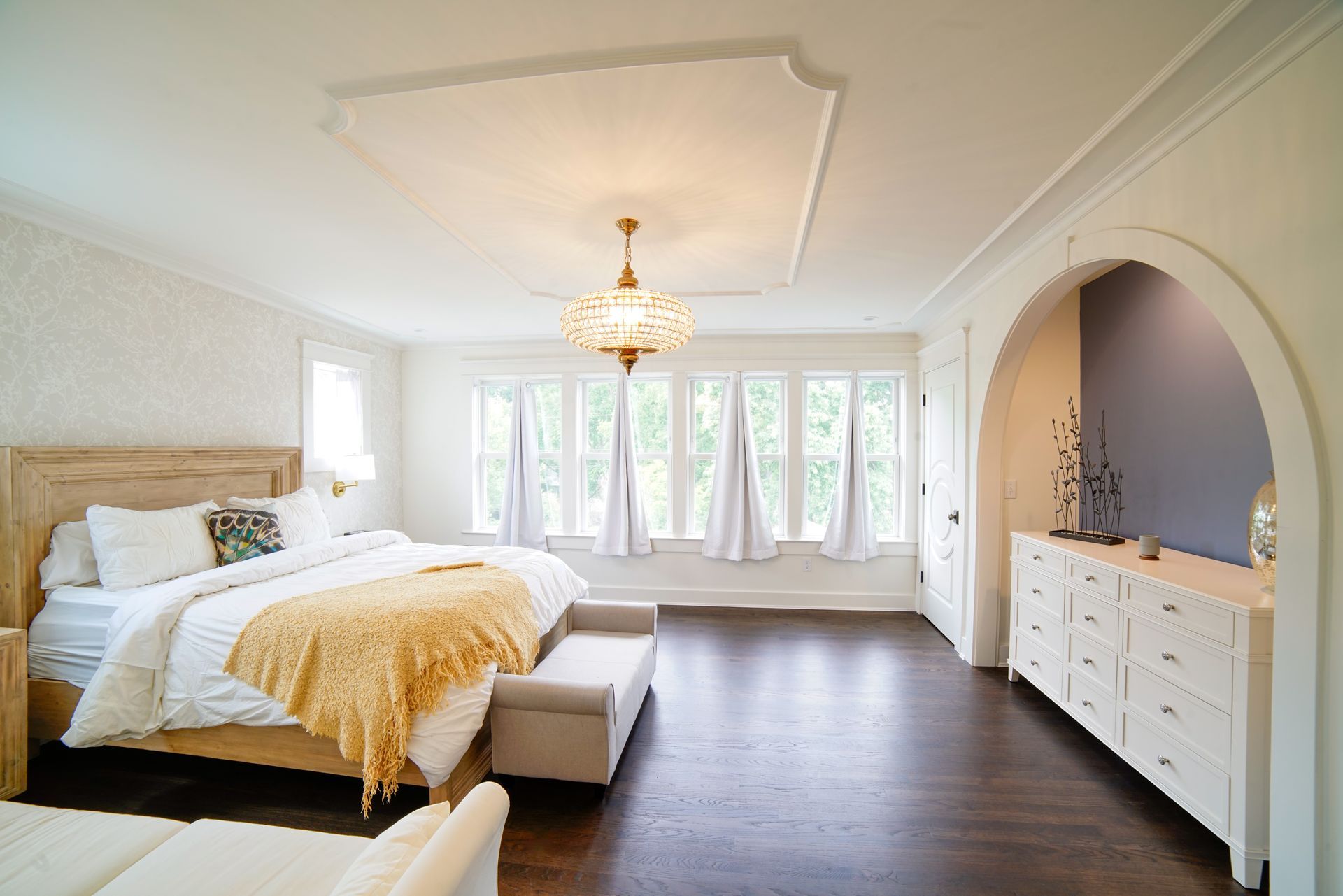 Bright bedroom with white walls, dark wood floor, chandelier, and dresser beneath an arched alcove