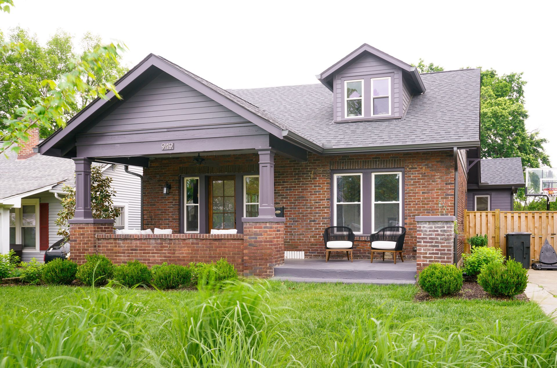 Brick bungalow with a dark gray upper facade and covered front porch, surrounded by green shrubs.