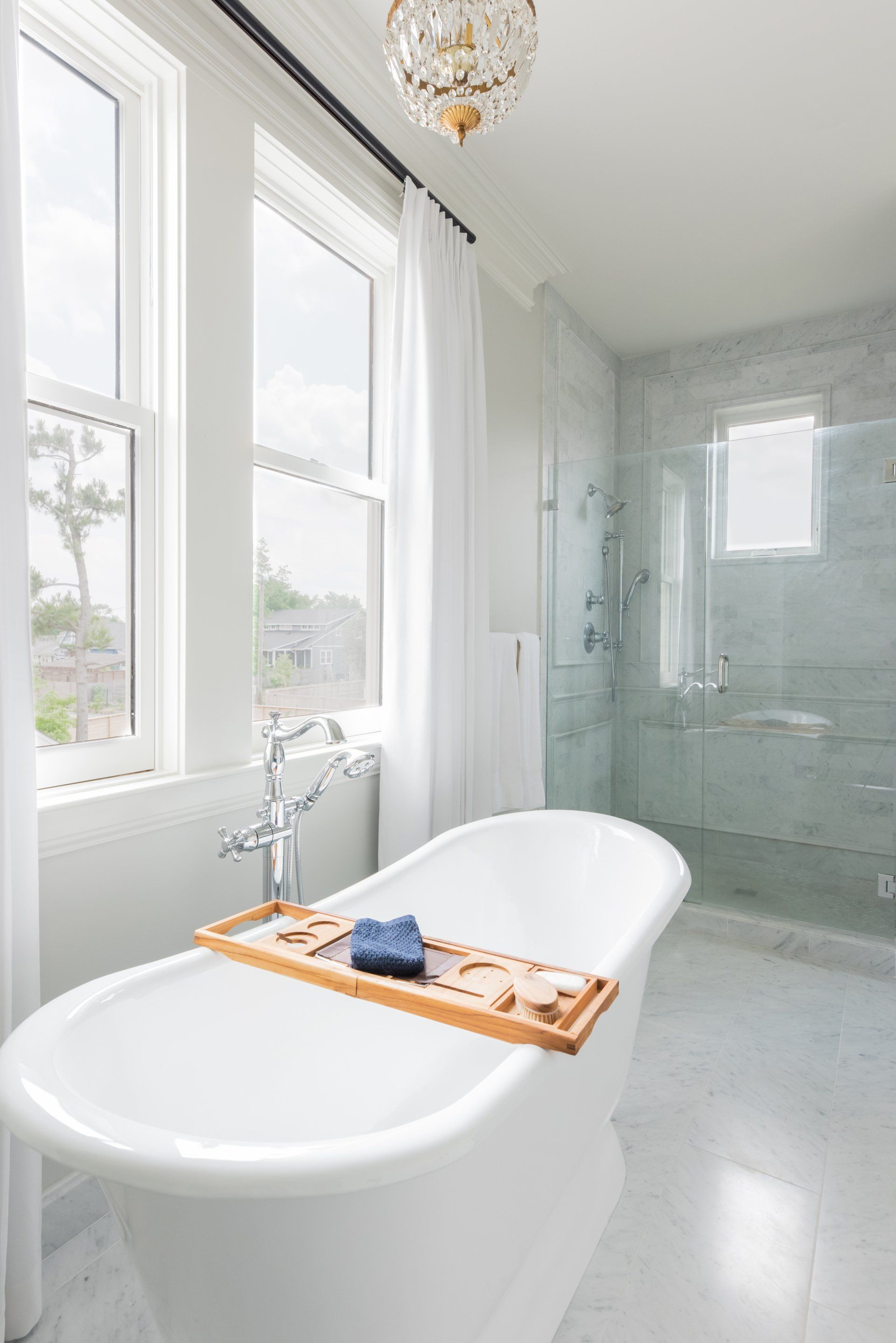 Bright white bathroom with a freestanding tub, tray, large windows, and glass-enclosed shower.