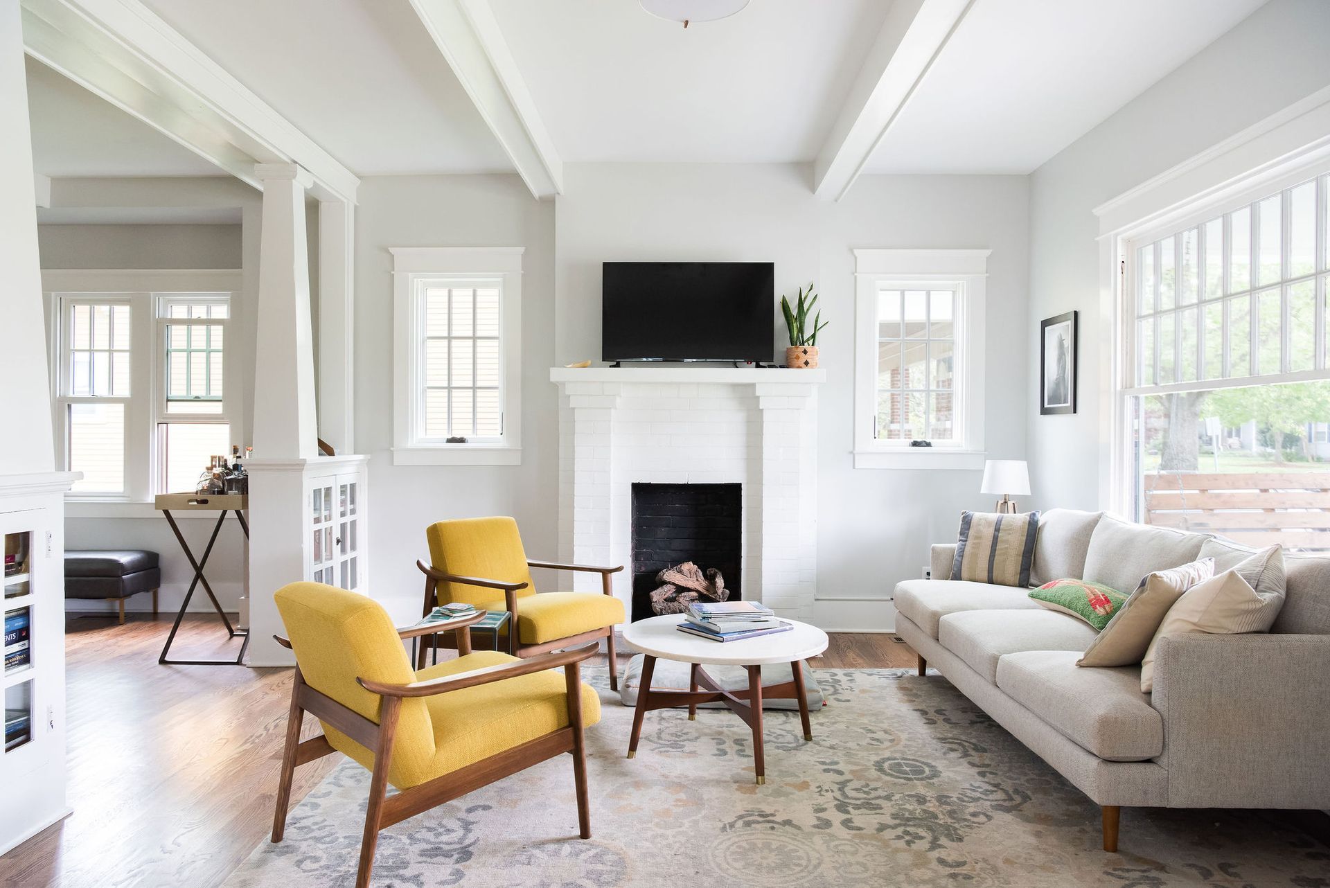 Bright living room with yellow chairs, gray sofa, fireplace, and TV, lit by large windows