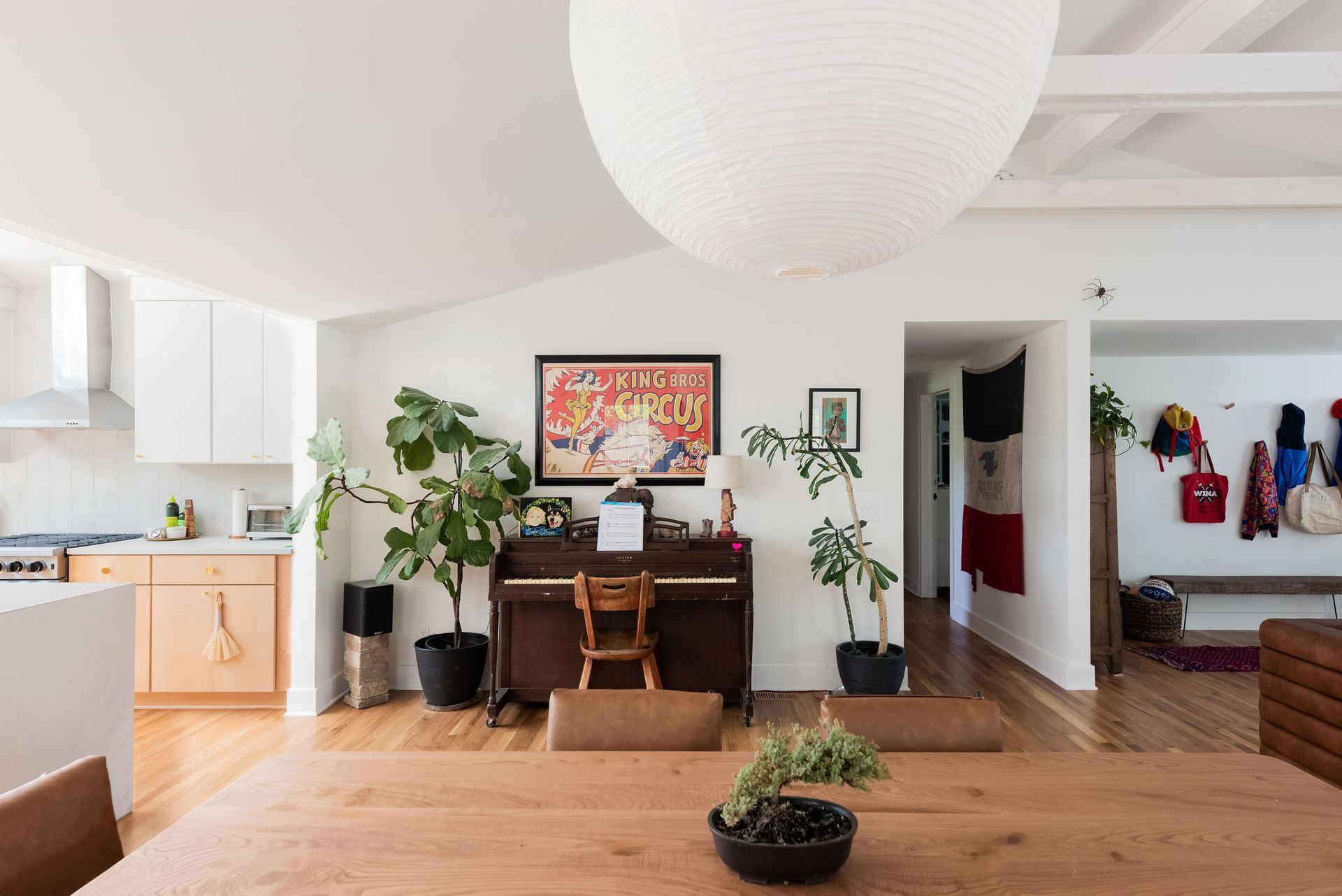 Bright living room with wooden table, piano, plants, and open kitchen in the background
