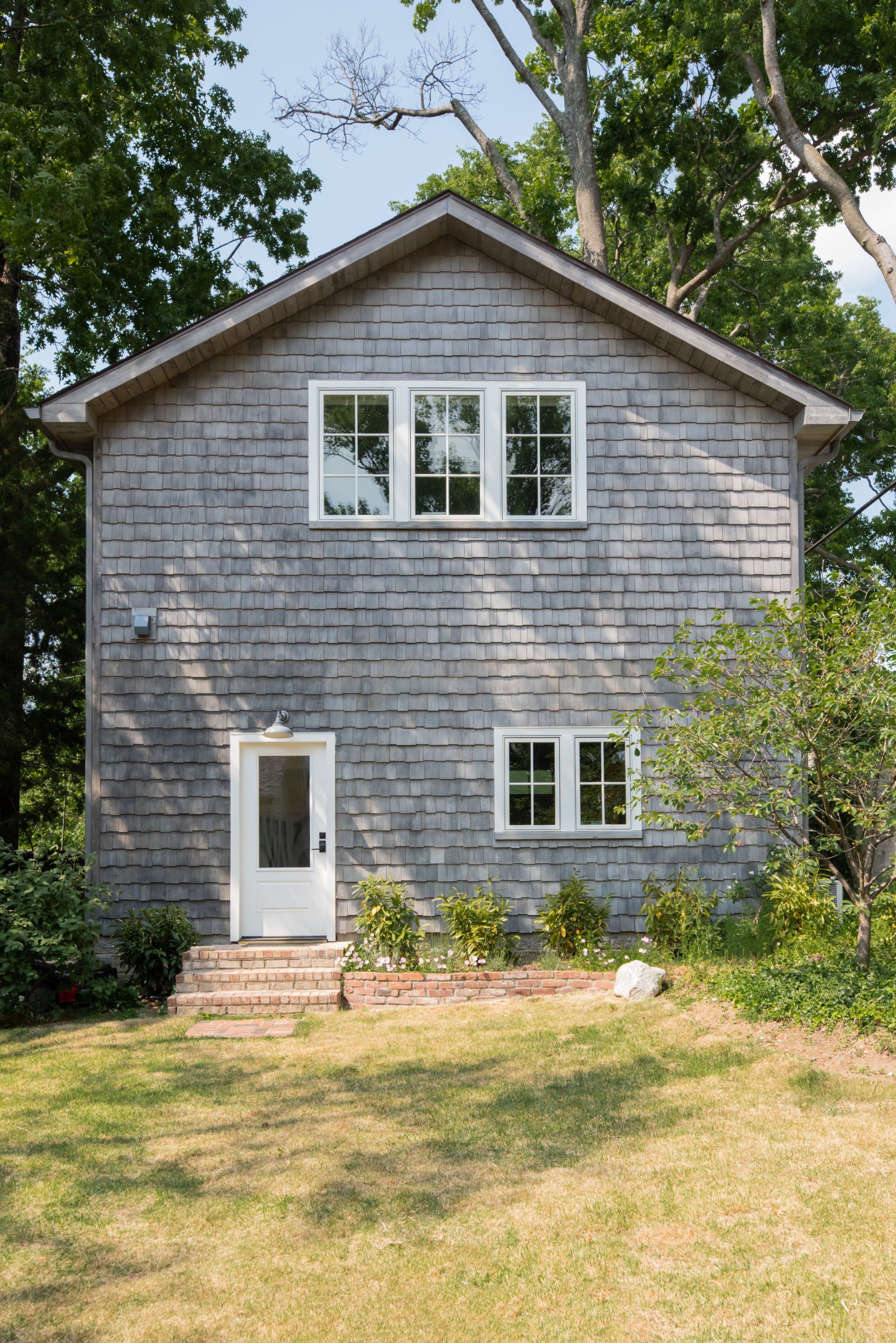 Gray shingled two-story house with white-trimmed windows and a small front door, surrounded by trees and yard