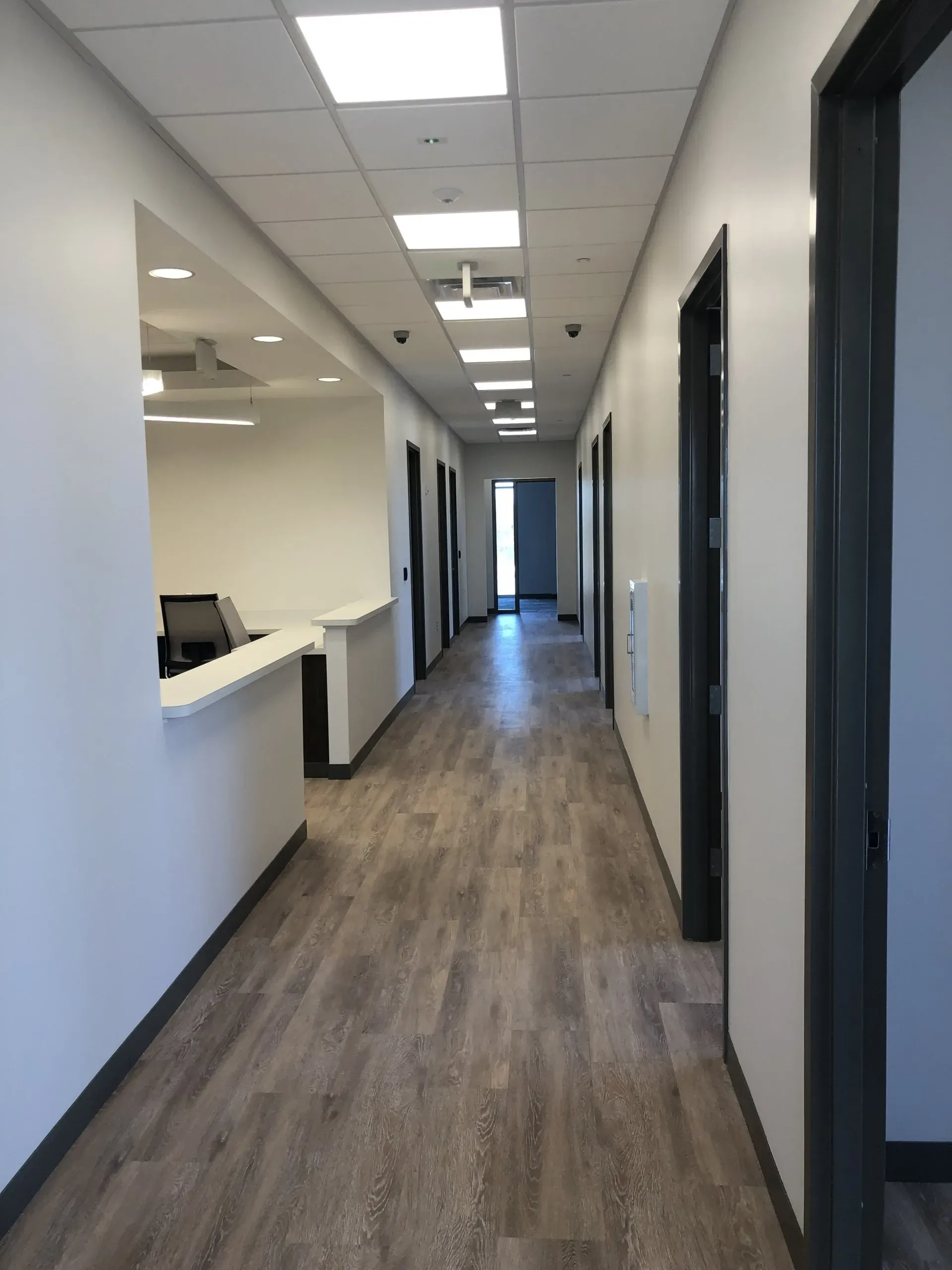 Empty office hallway with white walls, wood flooring, ceiling lights, and open doorways
