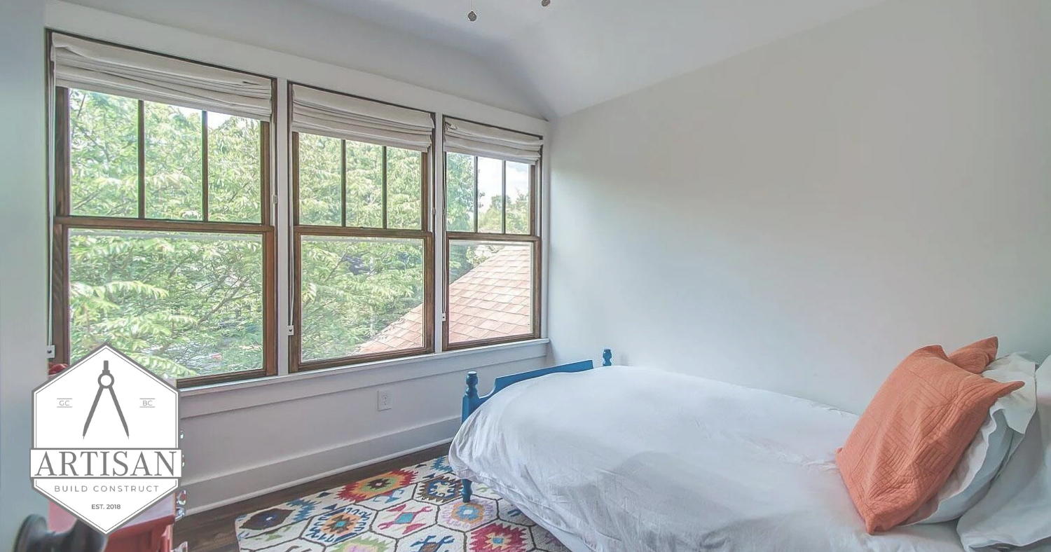 Bright bedroom with large windows, white bedding, coral pillow, and patterned rug.