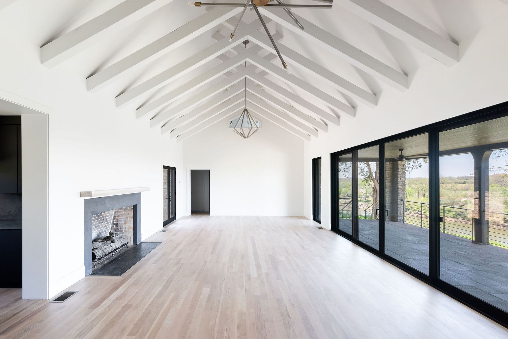 Bright empty living room with vaulted white ceiling, fireplace, light wood floors, and large glass doors to a patio