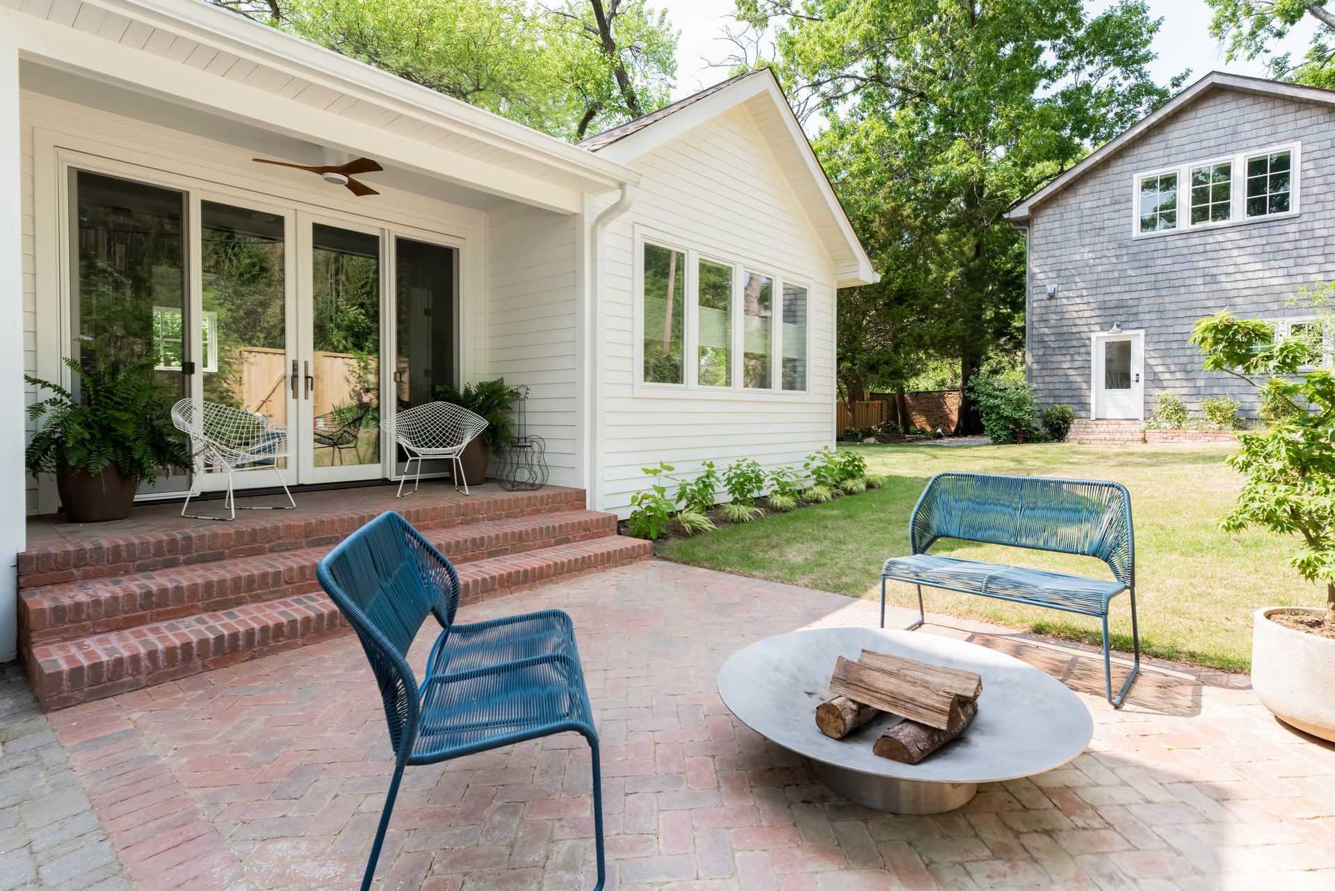 Patio with blue chairs and fire pit beside a white house with screened porch