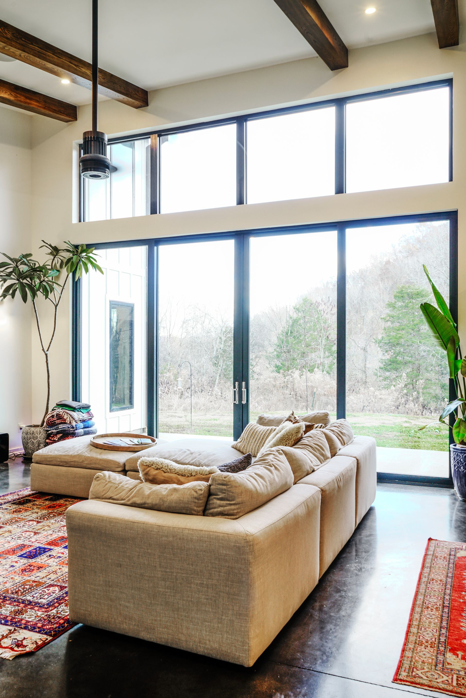 Bright living room with beige sofa, exposed beams, large windows, and natural light with trees outside