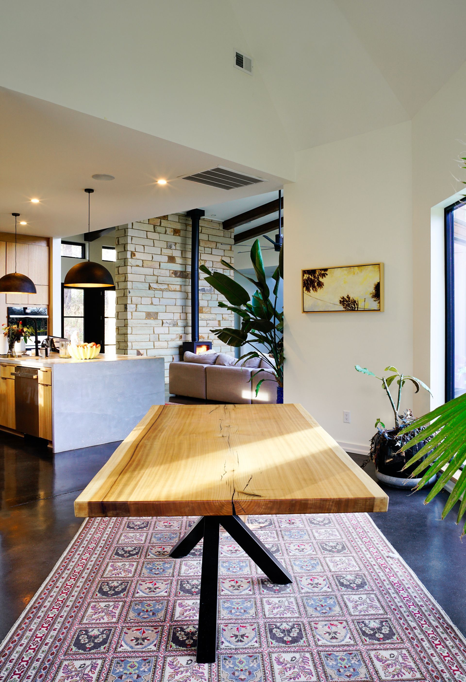 Modern dining area with a wooden table on a patterned rug, stone wall, and large window plants