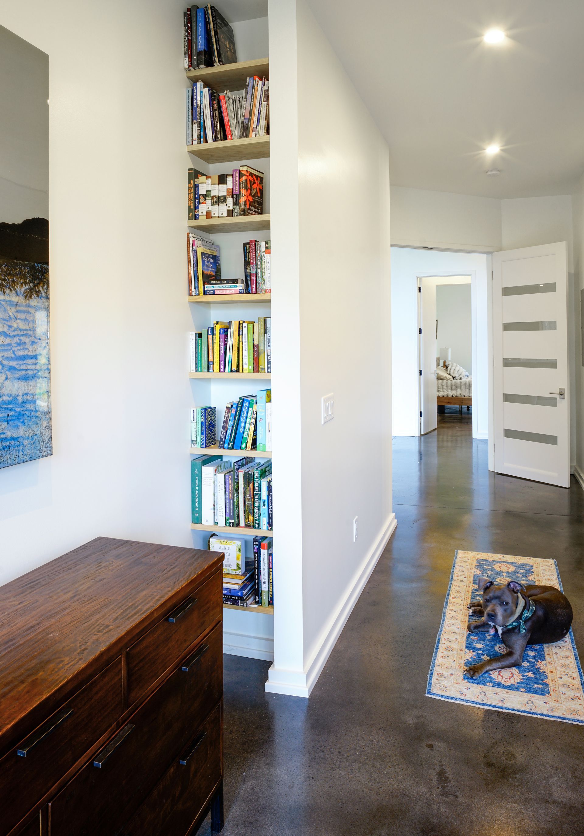 Narrow hallway with white walls, dark floor, bookshelf niche, and rug near an open door.