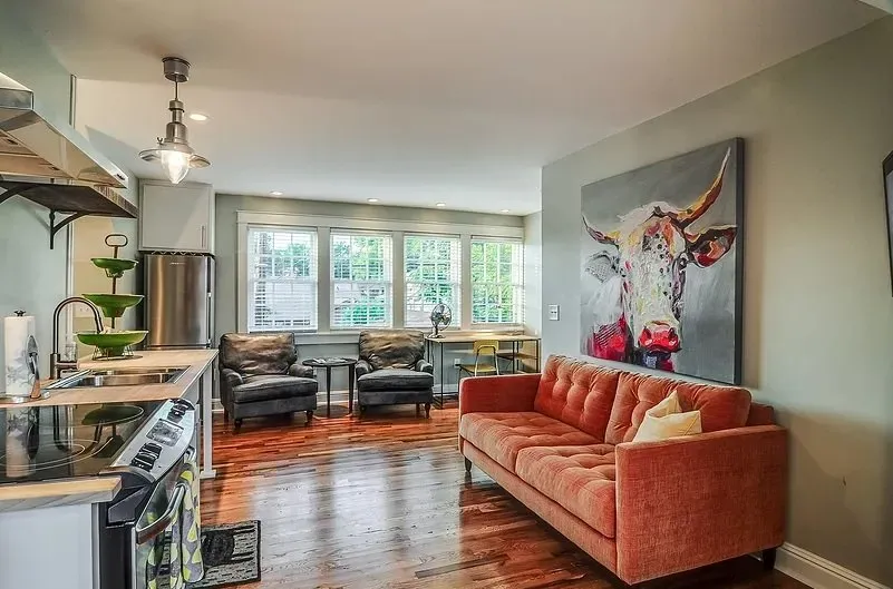 Bright living room with coral sofa, dark chairs, hardwood floors, and large windows beside cow artwork