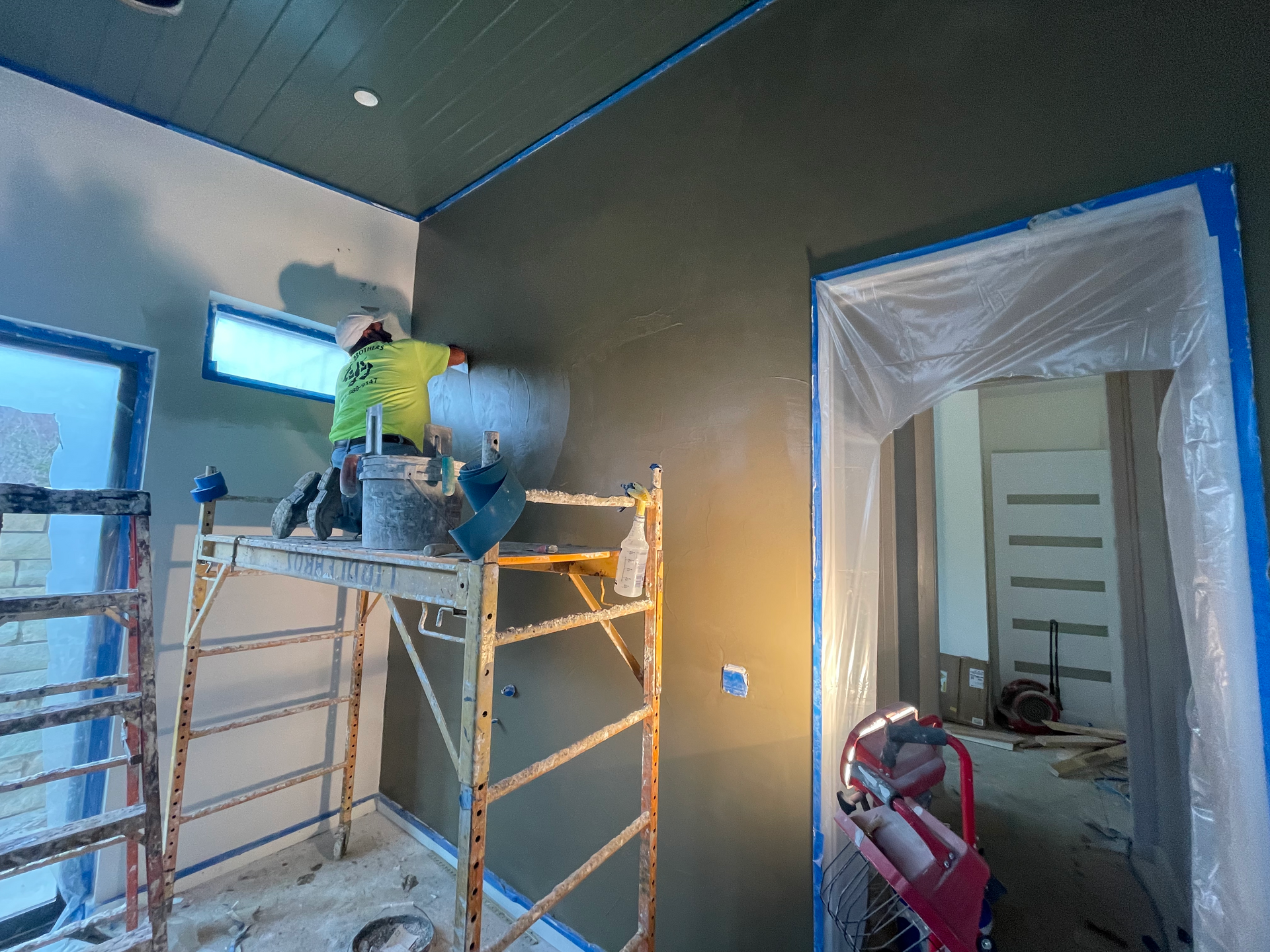 Worker on scaffolding painting a room interior with gray walls and blue tape around openings.