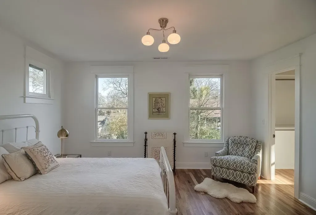 Bright bedroom with white bed, patterned chair, three windows, and a ceiling light fixture.