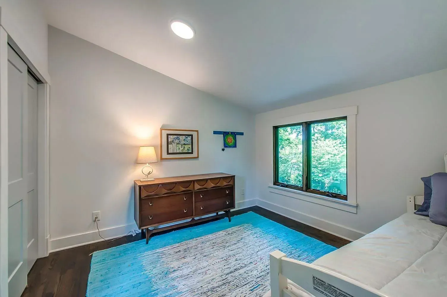 Bright bedroom with white walls, blue rug, wooden dresser, bed, and a window with green trees outside